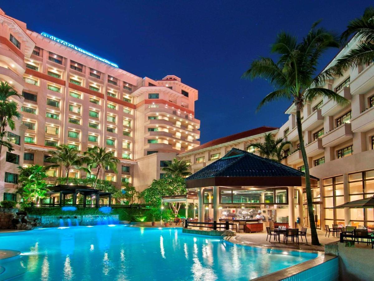 Paradox Singapore Merchant Court At Clarke Quay Exterior photo The photo depicts a luxurious hotel complex set against a twilight sky. In the foreground, there is a large swimming pool that reflects the light from surrounding lights and the sky. Lush greenery, including palm trees, adds to the tropical ambiance.