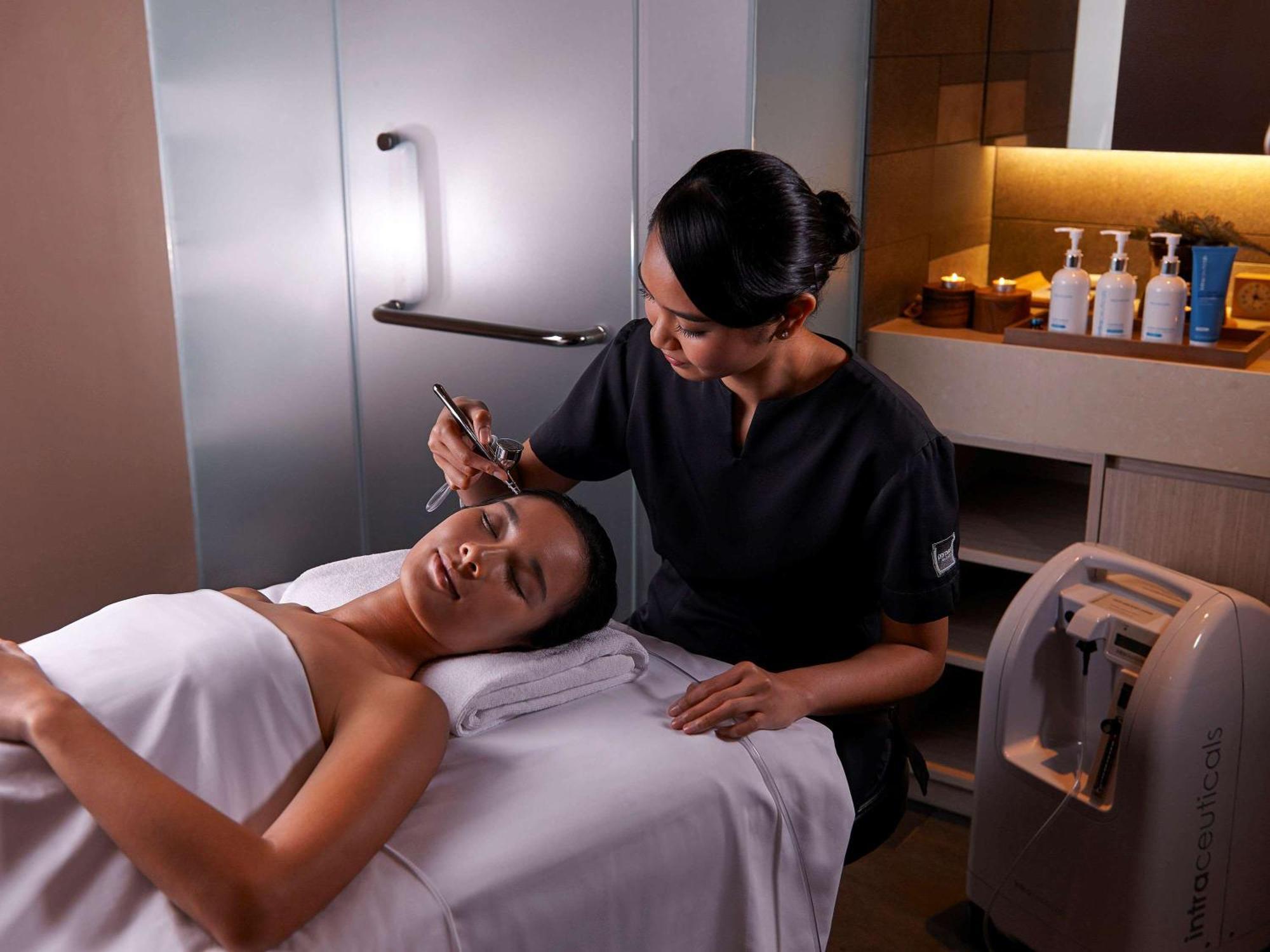 Paradox Singapore Merchant Court At Clarke Quay Exterior photo The photo shows a spa or beauty therapy setting where a woman is lying on a treatment table with a towel draped over her. A professional, dressed in a dark uniform, is performing a skincare treatment on her face. Various beauty products are visible o