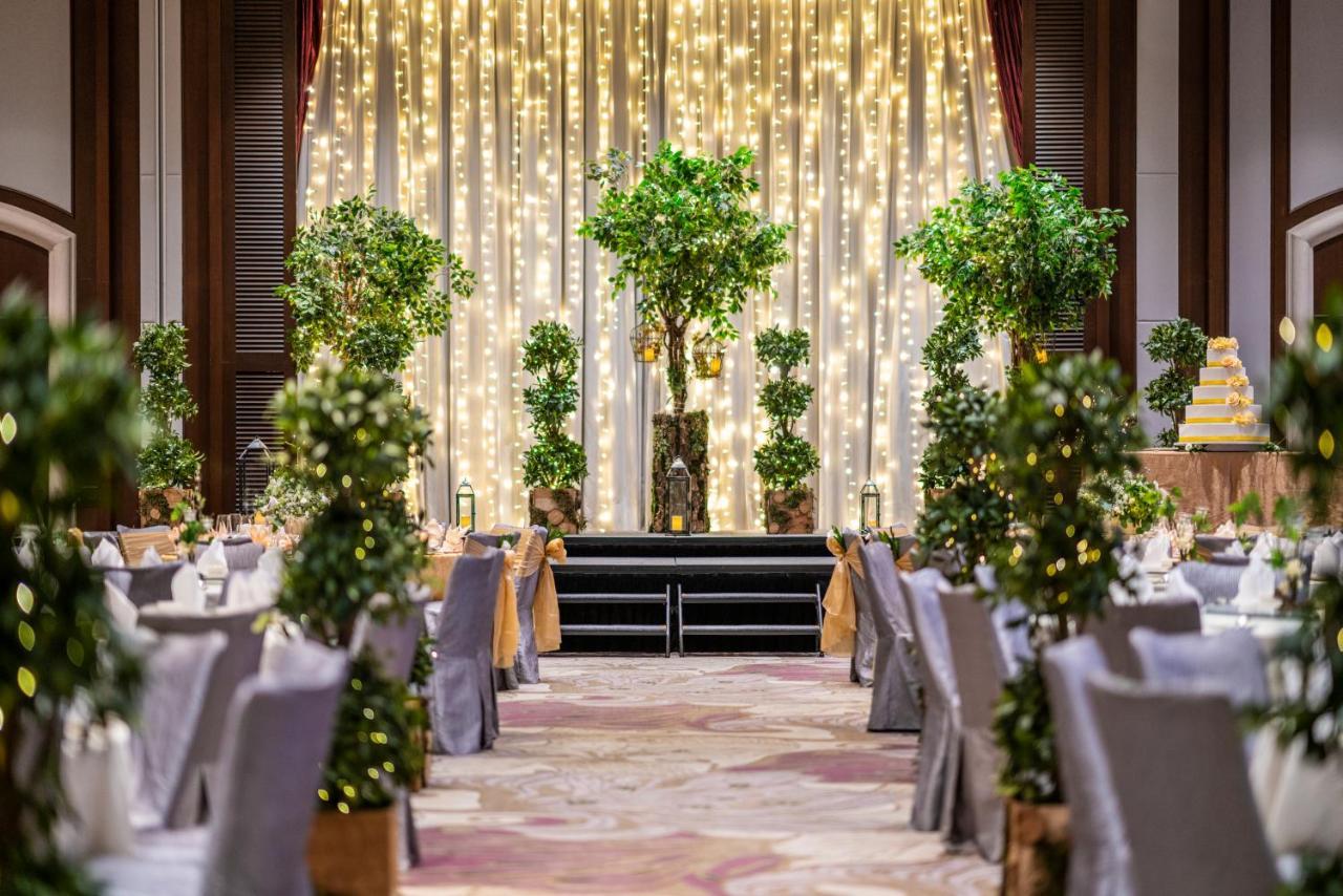 Paradox Singapore Merchant Court At Clarke Quay Exterior photo The image shows an elegantly decorated event space, likely for a wedding or formal celebration. In the center, there is a backdrop with draped fabric illuminated by soft lights, creating a warm ambiance. There are several decorative trees or potted p