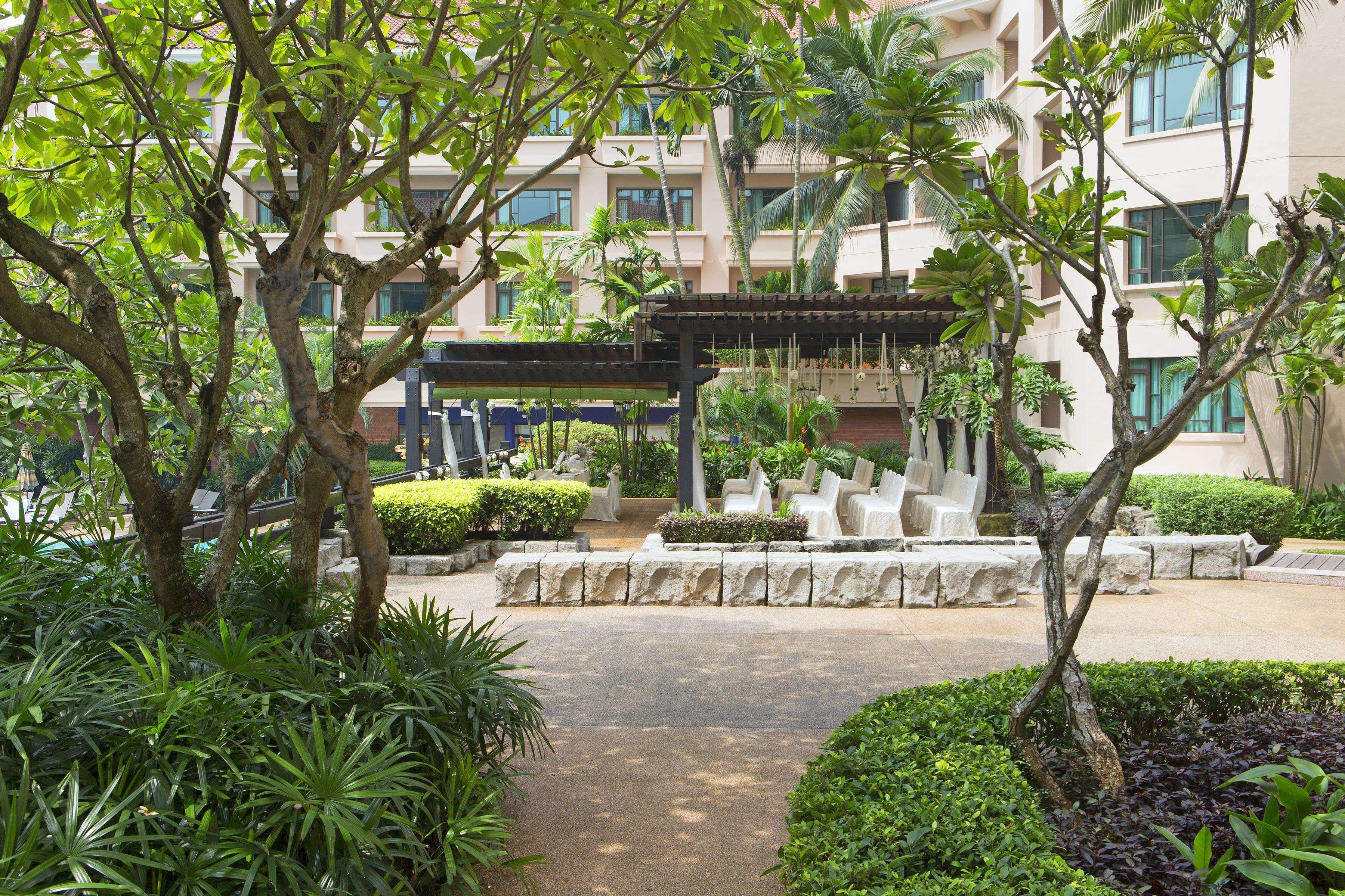 Paradox Singapore Merchant Court At Clarke Quay Exterior photo The photo depicts a garden or courtyard area surrounded by lush greenery. There are several trees and vibrant plants, creating a serene environment. In the center, there seems to be a stone seating arrangement under a covered structure, likely meant 