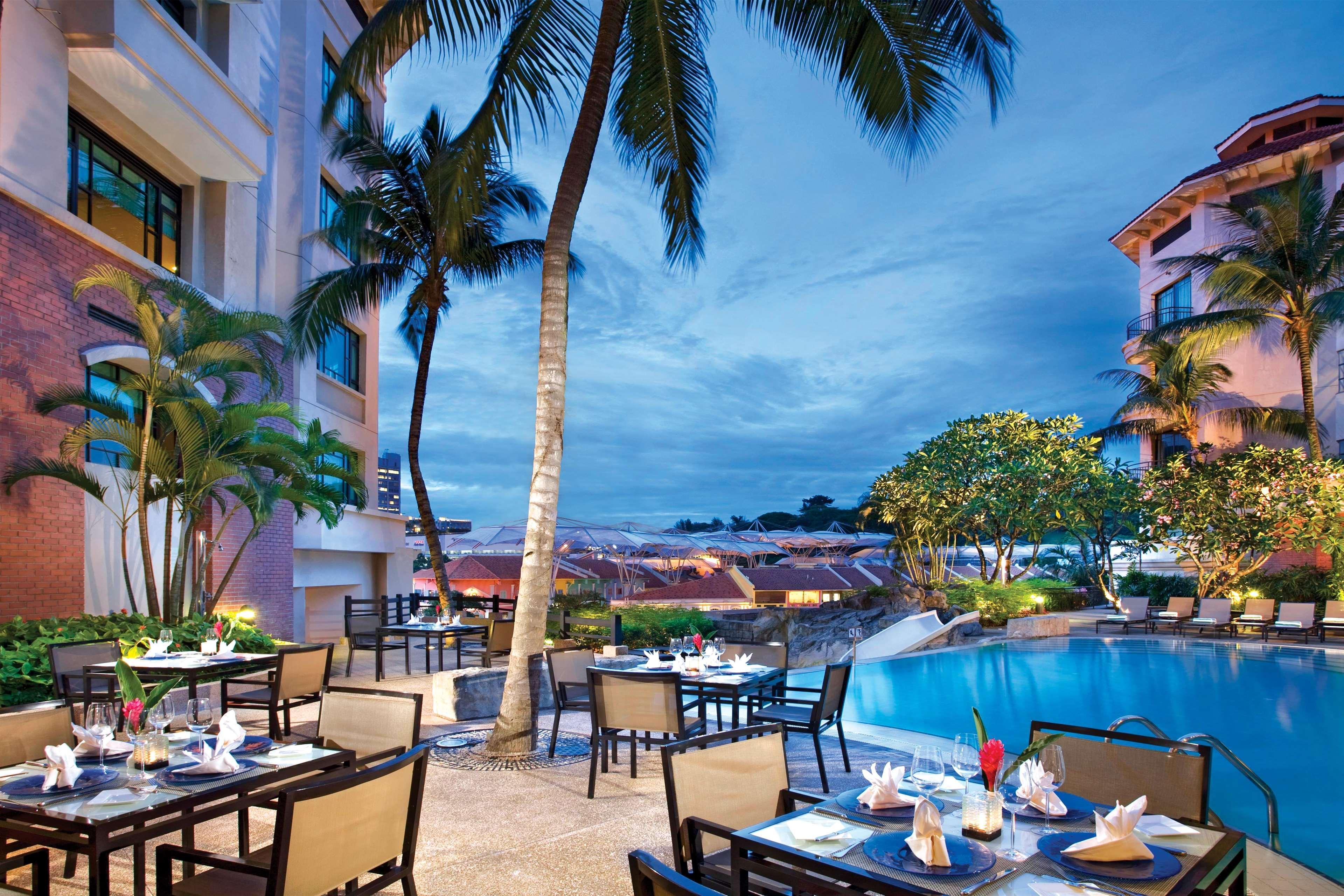 Paradox Singapore Merchant Court At Clarke Quay Restaurant photo The photo depicts a beautiful outdoor dining area next to a shimmering pool, set against a backdrop of palm trees and a dramatic evening sky. Tables are arranged elegantly with white tablecloths, chairs, and decorative napkins, suggesting a luxurious