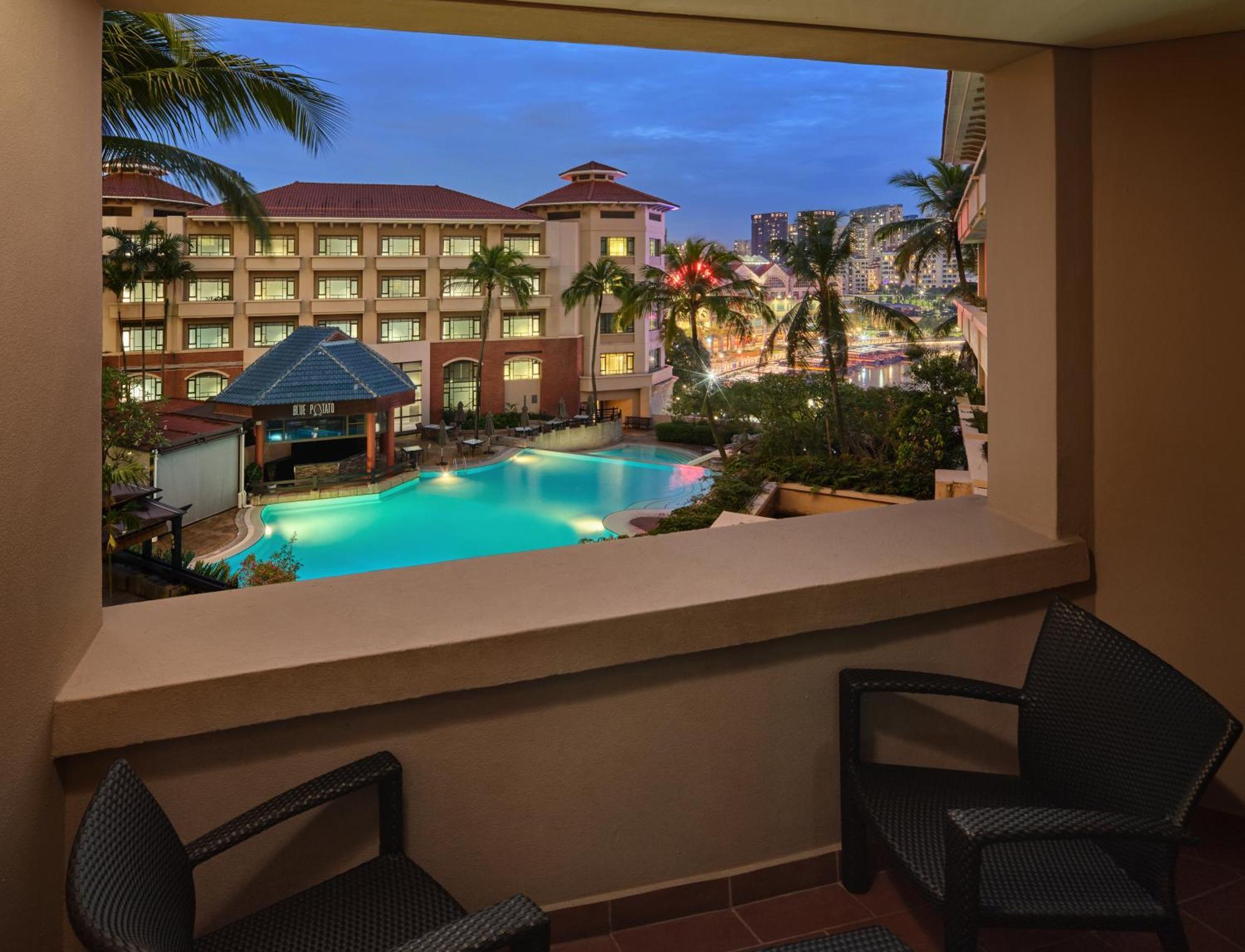 Paradox Singapore Merchant Court At Clarke Quay Exterior photo The image depicts a view from a balcony overlooking a pool area of a hotel or resort. The scene features palm trees surrounding the pool, which has a blue hue, possibly illuminated by lights. In the background, there are several buildings, likely par